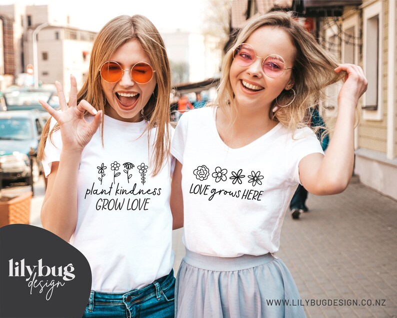 Op de afbeelding: Twee vrouwen dragen witte T-shirts met verschillende bloemenontwerpen en tekst. Het eerste shirt zegt "plant kindness GROW LOVE" en het tweede shirt zegt "LOVE grows HERE".