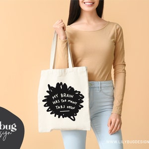 May include: A white tote bag with black text that reads "My brain has too many tabs open". The text is in a hand-drawn style and is surrounded by a black scribble.