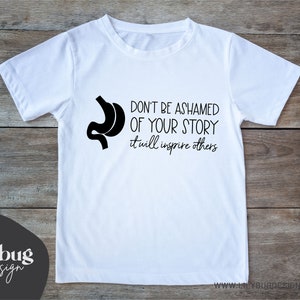 May include: White t-shirt with a black silhouette of a stomach and the text "Don't be ashamed of your story it will inspire others".