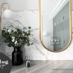 May include: A gold-framed oval mirror with the text "Happiness looks good on you, girl" reflected in the glass. A vase with green eucalyptus branches sits on a marble countertop.