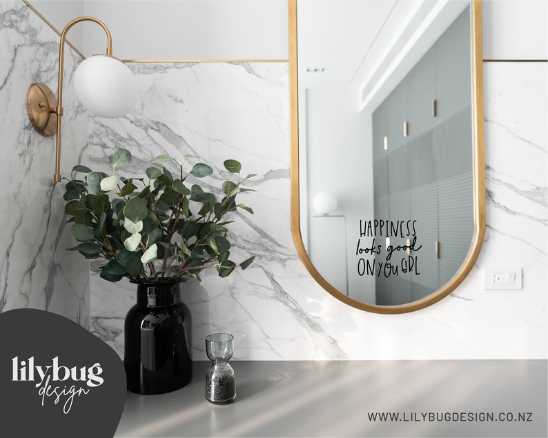 May include: A gold-framed oval mirror with the text "Happiness looks good on you, girl" reflected in the glass. A vase with green eucalyptus branches sits on a marble countertop.