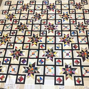 Może przedstawiać: Kołdra z geometrycznym wzorem czarnych kwadratów i gwiazd. Gwiazdy są wykonane z różnych tkanin w odcieniach brązu, czerwieni, błękitu i żółci. Kołdra jest wykonana z białej tkaniny o subtelnej fakturze.