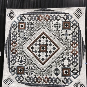 May include: A square quilt featuring a complex geometric pattern in black, white, gray, and copper tones. The design includes a central diamond, intricate border motifs, and four corner squares, all finished with detailed white machine quilting stitches.