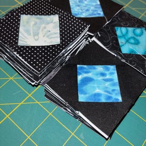 May include: Stacks of small, square fabric pieces for quilting. The squares feature a black background with white polka dots or swirls, and a central square with a blue water pattern or a white design. The fabric is stacked on a green cutting mat.