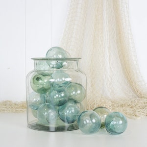May include: A clear glass jar filled with multiple blue-green glass fishing floats. Some floats are outside the jar. A beige fishing net hangs in the background. The jar is on a white surface.