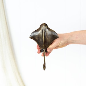 May include: A bronze-coloured wall hook in the shape of a manta ray. The hook is held in a hand against a white backdrop. The ray has a textured surface and a single hook at the bottom. The hook is made of metal and is designed for hanging items.