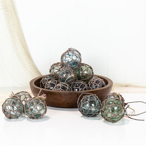 May include: A wooden bowl filled with a collection of glass fishing floats, each wrapped in brown twine. The floats are various shades of blue and green. Several floats are also scattered on a white surface.
