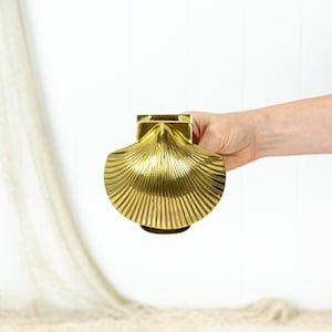 May include: A gold-coloured, seashell-shaped decorative object. The object has a ridged, fan-like design and a square top. The object is held by a person's hand against a white background.