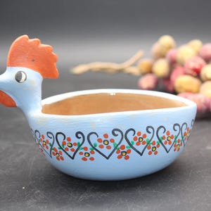 May include: A light blue ceramic bowl shaped like a rooster. The bowl has a brown interior and is decorated with a floral pattern in red, green, and black. The rooster has an orange comb and wattle.