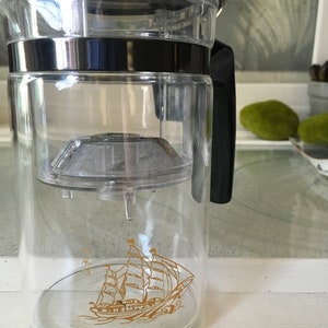 May include: Clear glass tea infuser with a black handle and lid. The infuser has a gold sailboat design on the front. The tea infuser has a stainless steel mesh filter inside.