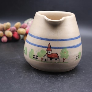 May include: A cream-colored ceramic pitcher with a blue and white stripe design. The pitcher features a hand-painted scene of a small church with a red roof, green trees, and a fence.