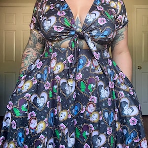 May include: A black dress with a V-neck and tie detail. The dress is patterned with heart-shaped portraits, pink flowers, and white stars. The short-sleeved dress has a full skirt.