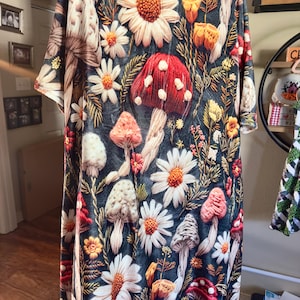 May include: A dark blue, short-sleeved tunic featuring a repeating pattern of mushrooms and daisies in shades of red, white, orange, and beige. The floral design is set against a dark background, creating a whimsical and nature-inspired aesthetic.