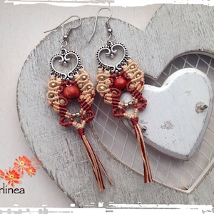 Peut inclure: Une paire de boucles d'oreilles faites à la main avec un motif en forme de cœur. Les boucles d'oreilles sont faites de cordon rouge et beige et présentent une petite perle rouge au centre de chaque cœur. Les boucles d'oreilles sont suspendues à des crochets argentés.