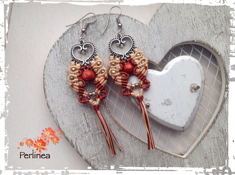 Puede incluir: Un par de pendientes hechos a mano con un dise&ntilde;o en forma de coraz&oacute;n. Los pendientes est&aacute;n hechos con cord&oacute;n rojo y beige y presentan una peque&ntilde;a cuenta roja en el centro de cada coraz&oacute;n. Los pendientes cuelgan de ganchos plateados.