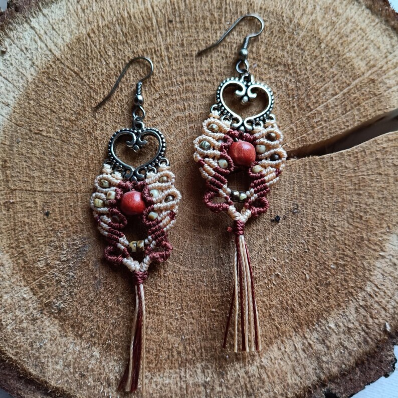 Puede incluir: Un par de pendientes hechos a mano con un dise&ntilde;o en forma de coraz&oacute;n. Los pendientes est&aacute;n hechos con cord&oacute;n de macram&eacute; marr&oacute;n y beige y presentan una cuenta de madera roja en el centro. Los pendientes est&aacute;n terminados con un dije de coraz&oacute;n plateado y una borla.