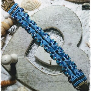 Puede incluir: Un brazalete azul y negro con cuentas y un cierre plateado. El brazalete est&aacute; sobre un fondo de madera blanco en forma de coraz&oacute;n con conchas.