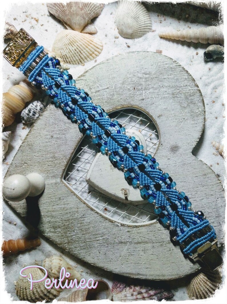 Puede incluir: Un brazalete azul y negro con cuentas y un cierre plateado. El brazalete est&aacute; sobre un fondo de madera blanco en forma de coraz&oacute;n con conchas.