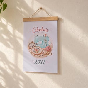May include: A wall calendar for 2027 featuring the text Calendario and an illustration of a light blue sewing machine, an embroidery hoop with flowers, a ball of pink yarn, a pair of scissors, and a small coin purse, suspended by a wooden frame.