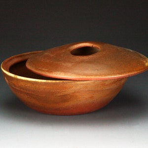 May include: A brown ceramic bowl with a lid. The bowl has a slightly rounded shape and the lid has a small hole in the center. The bowl and lid have a rustic, earthy look.