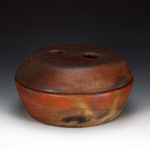 May include: A handmade, ceramic lidded bowl with a rustic, earthy aesthetic. The bowl is primarily reddish-brown with areas of orange and black, and the lid features two small holes. The piece has a rounded shape and a matte finish.