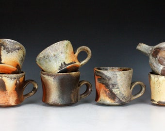 May include: Six ceramic mugs with a variety of brown, orange, and white glaze patterns. The mugs are stacked in pairs, with one mug on top of another. The mugs have a rustic, handmade look.