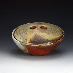 May include: A round ceramic bowl with a matching lid featuring two circular openings. The exterior displays a mottled glaze with blended tones of deep red, earthy brown, and muted gray, creating a textured appearance across the entire surface.