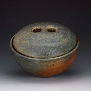May include: A round ceramic lidded bowl with a textured, mottled surface in shades of charcoal gray, muted green, and burnt orange. The lid features two circular openings at the top, and the bowl has a curved, deep basin shape with a rustic finish.
