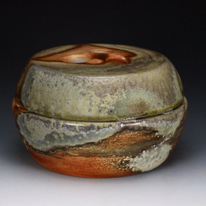 May include: A handcrafted ceramic lidded jar with a rounded shape. The jar features a blend of earthy tones, including shades of brown, orange, and green. The lid has a unique, organic design, and the overall aesthetic is rustic and artistic.