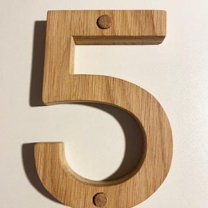 May include: A wooden number five, made from light oak. The number is thick and three-dimensional, with visible wood grain. Two small wooden pegs secure the number. Suitable for home decor or as a house number.