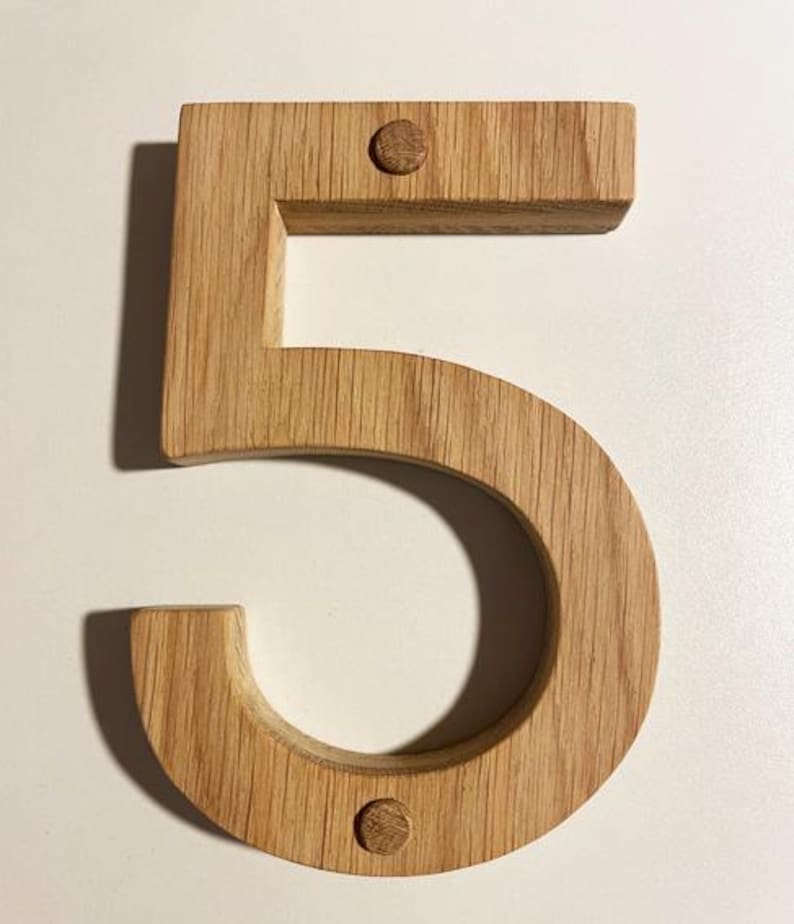 May include: A wooden number five, made from light oak. The number is thick and three-dimensional, with visible wood grain. Two small wooden pegs secure the number. Suitable for home decor or as a house number.