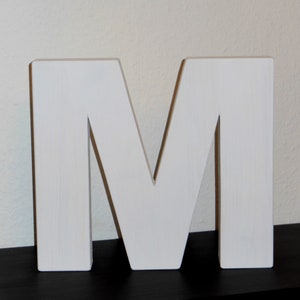 May include: A large, white wooden letter "M" stands on a black surface against a neutral wall. The letter is painted white and has a blocky, three-dimensional design, suitable for home decor or personalized gifts.