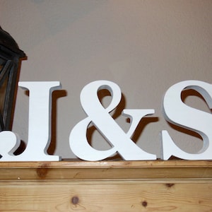 Op de afbeelding: Witte houten letters "J & S" worden weergegeven op een houten oppervlak. De letters zijn dik en staan rechtop, met een schaduweffect. Een donkere houten lantaarn is gedeeltelijk zichtbaar aan de linkerkant. De achtergrond is een neutrale muur.
