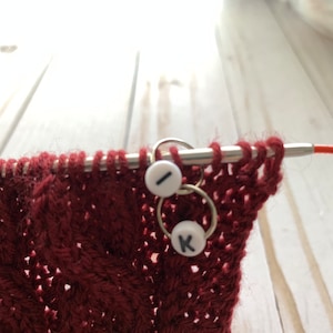 May include: Close-up of a burgundy knitted fabric with a cable pattern, held on knitting needles. Two stitch markers with white beads and black letters "I" and "K" are attached to the needles, indicating the pattern's progress. The needles are silver.