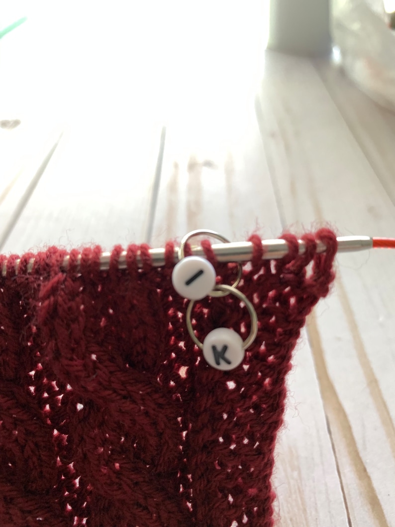 May include: Close-up of a burgundy knitted fabric with a cable pattern, held on knitting needles. Two stitch markers with white beads and black letters "I" and "K" are attached to the needles, indicating the pattern's progress. The needles are silver.