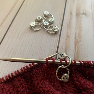 May include: Close-up of a red knitted fabric with a silver knitting needle and two silver stitch markers with the letter "K" on them.