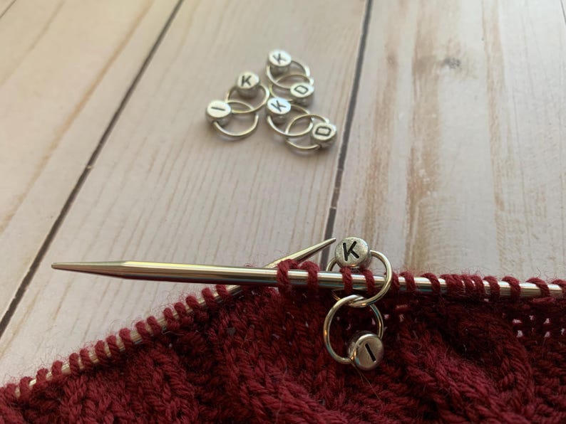 May include: Close-up of a red knitted fabric with a silver knitting needle and two silver stitch markers with the letter "K" on them.