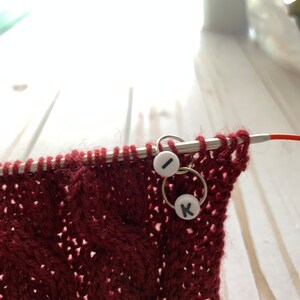 May include: Close-up of a burgundy knitted fabric with a cable pattern, held on a metal knitting needle. Two stitch markers with the letters "I" and "K" are attached to the needle. The background is blurred.