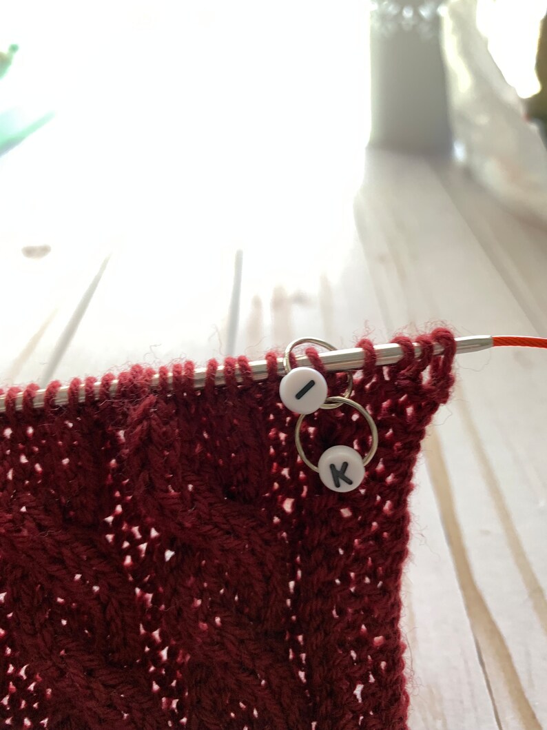 May include: Close-up of a burgundy knitted fabric with a cable pattern, held on a metal knitting needle. Two stitch markers with the letters "I" and "K" are attached to the needle. The background is blurred.