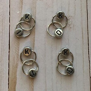 May include: Four sets of silver-toned, double-loop stitch markers with black letter beads. Each set has a letter 'K' and a blank bead.