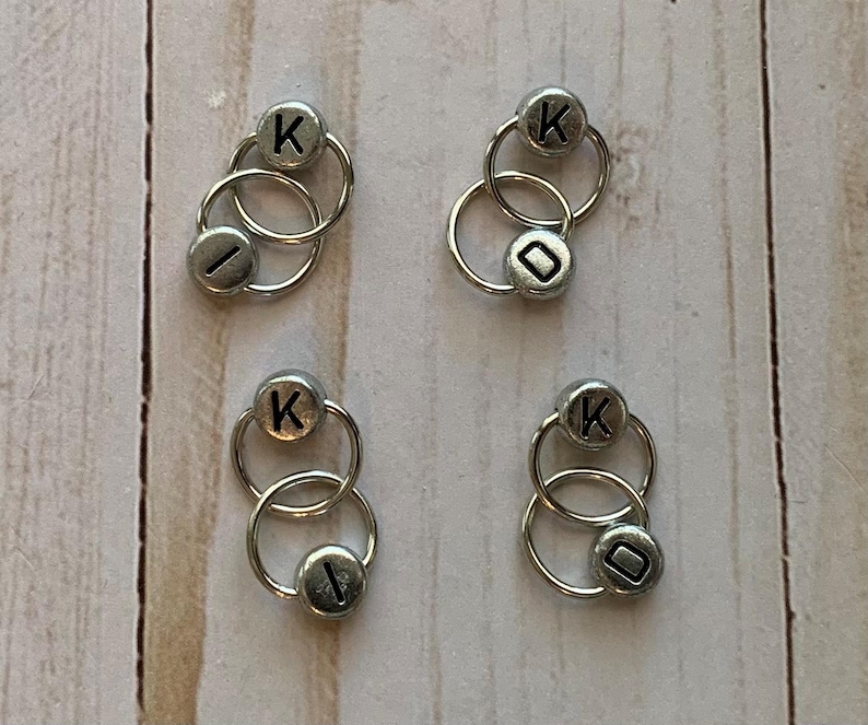 May include: Four sets of silver-toned, double-loop stitch markers with black letter beads. Each set has a letter 'K' and a blank bead.