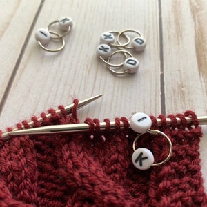 May include: Close-up of knitting needles with a burgundy knitted fabric and stitch markers. The stitch markers are small white beads with black letters, attached to gold-colored rings. The needles are silver, and the fabric has a textured pattern.
