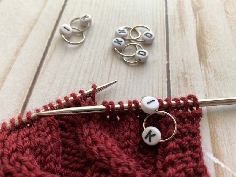 May include: Close-up of knitting needles with a burgundy knitted fabric and stitch markers. The stitch markers are small white beads with black letters, attached to gold-colored rings. The needles are silver, and the fabric has a textured pattern.