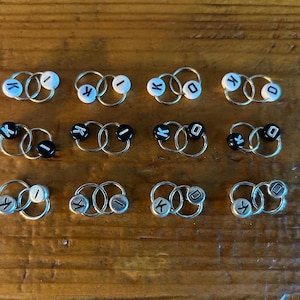 May include: A selection of small hoop earrings in silver. Each earring has two linked hoops and a small, round bead with a letter. The beads are either white with blue letters or black with white letters.