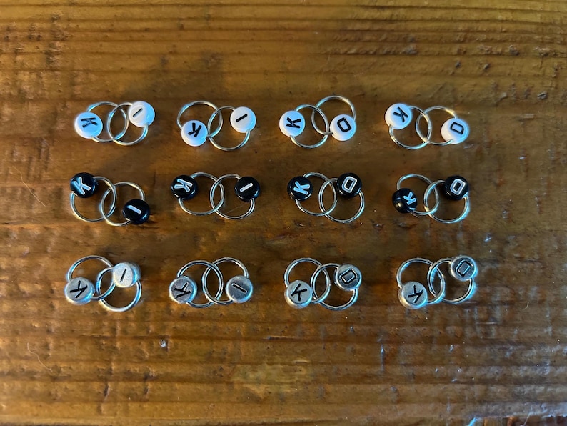 May include: A selection of small hoop earrings in silver. Each earring has two linked hoops and a small, round bead with a letter. The beads are either white with blue letters or black with white letters.