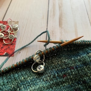 May include: Close-up of a green and blue knitted fabric with two silver stitch markers labeled "I" and "K". The fabric is being worked on with wooden knitting needles.