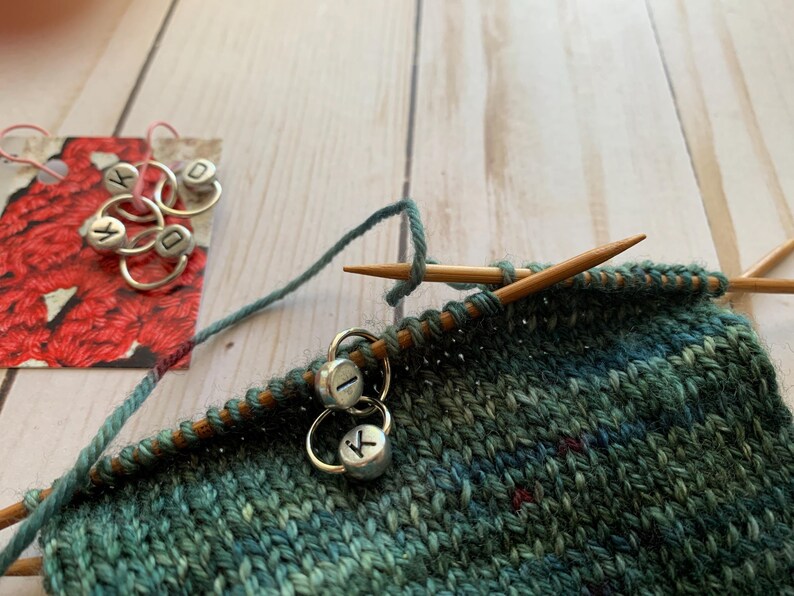 May include: Close-up of a green and blue knitted fabric with two silver stitch markers labeled "I" and "K". The fabric is being worked on with wooden knitting needles.
