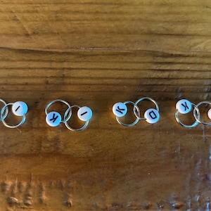 May include: Four sets of small, silver rings, each with two interlocked rings. Each set has a small, round, white bead with a black letter. The letters spell "KIKO". The background is a wooden surface.