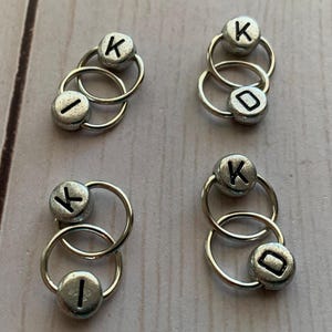 May include: Four sets of silver-toned metal ring stitch markers with black letter beads. Each set has two rings and two beads. The beads are labelled with the letter "K" and the letter "I".