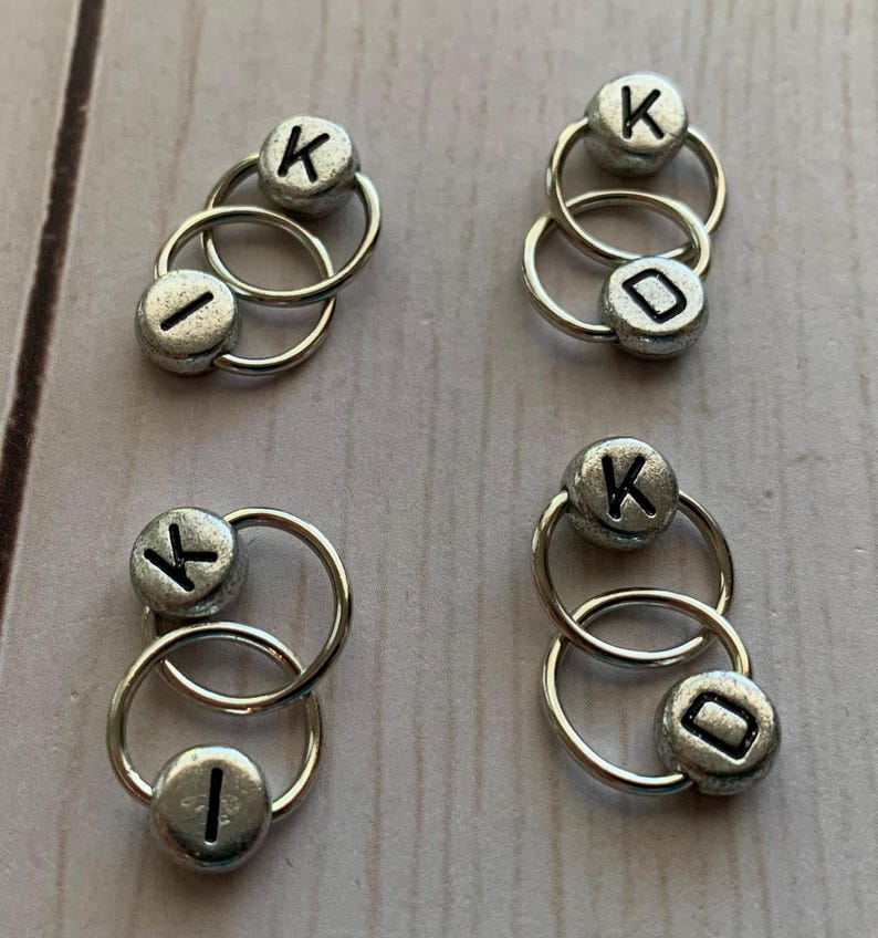 May include: Four sets of silver-toned metal ring stitch markers with black letter beads. Each set has two rings and two beads. The beads are labeled with the letter "K" and the letter "I".